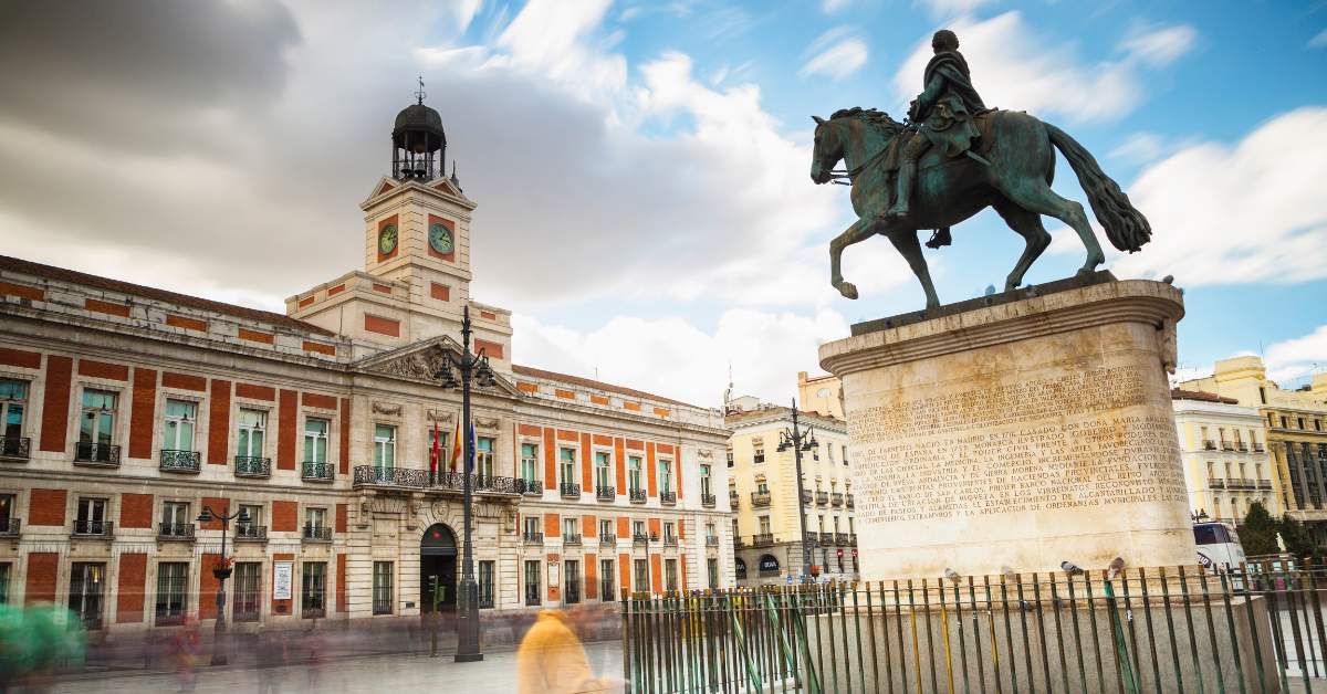 A daytime, long-exposure photograph of the equestrian statue of King Charles III in Madrid's bustling Puerta del Sol square, with the historic Real Casa de Correos building and its iconic clock tower in the background under a blue, cloudy sky.